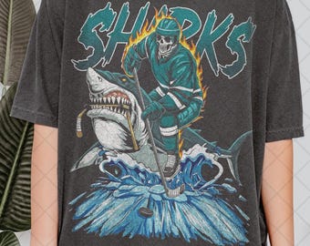 May include: Dark gray t-shirt with a graphic of a skeleton hockey player riding a shark, with the word "SHARKS" in teal. The hockey player is wearing teal and white hockey gear. The shark has a hockey stick in its mouth.