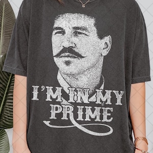 May include: Dark gray t-shirt featuring a distressed black and white portrait of a man with a large mustache. The text "I'M IN MY PRIME" is printed in white below the portrait. The shirt has short sleeves and a crew neck.