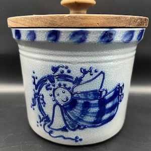 May include: A blue and white ceramic cookie jar with a wooden lid. The jar features a blue and white illustration of an angel with a halo and wings.