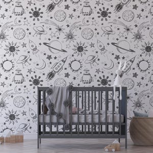 May include: Black and white space-themed wallpaper with a repeating pattern of stars, planets, rockets, and UFOs. The wallpaper is shown in a nursery setting with a crib and a stuffed animal.