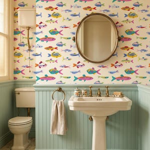 Puede incluir: Un baño con papel tapiz de patrón de peces coloridos, un lavabo de pedestal blanco con grifería de latón, un espejo redondo, una toalla beige en un anillo, un inodoro blanco y paneles de pared verde claro sobre un suelo de baldosas hexagonales.