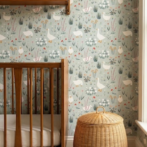 Op de afbeelding: Een kinderkamer met een houten wieg, een rieten mand en een plank met beeldjes. Het behang heeft een patroon van witte ganzen, bloemen en groen op een lichtgroene achtergrond. Een lamp met een beige kap staat op de plank.