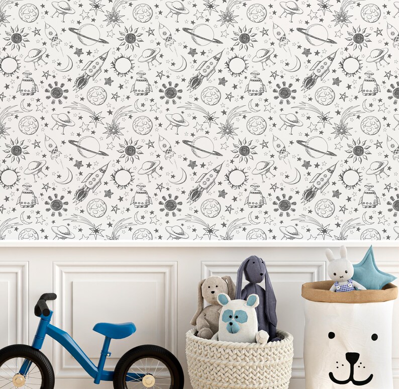 May include: A white wall with a black and white space-themed wallpaper featuring rockets, planets, stars, and suns. A blue children's bicycle is in the foreground, and a basket of stuffed animals is in the lower right corner.
