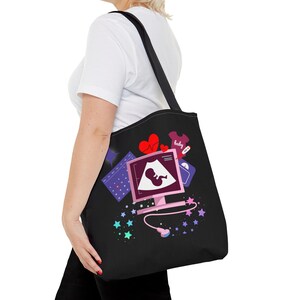 Peut inclure: Sac fourre-tout noir avec un motif d'écran d'ordinateur rose affichant une échographie de bébé. Le motif comprend des cœurs, un body de bébé, une balance, un calendrier et des étoiles colorées. Le sac est porté à l'épaule.