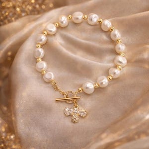 May include: A gold-toned bracelet featuring a strand of white pearls and gold beads. The bracelet has a decorative clasp and a charm with clear crystals. The jewelry is displayed on a gold satin fabric.