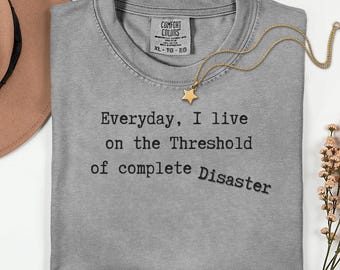 May include: A gray Comfort Colors t-shirt with the text "Everyday, I live on the Threshold of complete Disaster" in black. A gold star pendant necklace is draped across the shirt. A brown hat and flowers are in the background.