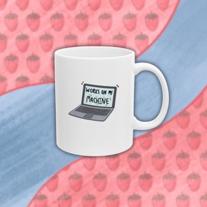 以下が含まれることがあります： 黒いラップトップのイラストと「WORKS ON MY MACHINE」という黒いテキストが描かれた白いセラミックマグカップ