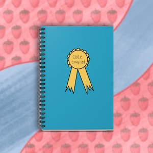 Pode incluir: Um caderno espiral azul com uma fita amarela e um círculo amarelo com o texto "código compilado" nele.