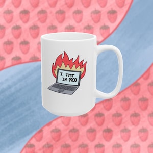 May include: White ceramic mug with a black laptop on fire with the text "I test in prod" on the screen. The mug is on a pink and blue background with a strawberry pattern.