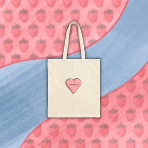 Op de afbeelding: Een beige tote bag met een roze hart waarop "<code />" staat. De tas staat voor een achtergrond van roze aardbeien.