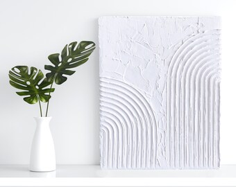 May include: White textured canvas painting with a raised design of white lines forming a curved pattern. The painting is on a white shelf with a green plant in a white vase.