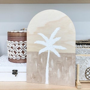 Puede incluir: Una palmera pintada de blanco sobre un panel de madera con fondo marrón y beige. El panel tiene forma de arco.