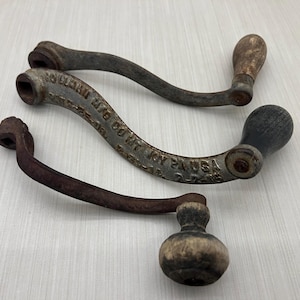以下が含まれることがあります： 木製のグリップが付いた、3つのアンティークの金属製クランクハンドル。ハンドルは湾曲しており、錆びた外観で、経年劣化の兆候が見られます。1つのハンドルには「JOY PA USA」の文字とその他の文字があります。