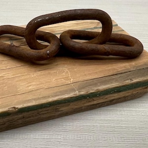 Puede incluir: Un bloque de madera rústica con tres eslabones de cadena de metal oxidado. La madera presenta vetas visibles y un acento verde en el borde. Los eslabones de la cadena son de forma ovalada. El bloque mide unos 15 cm de largo.