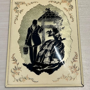 May include: A framed silhouette artwork depicting a couple standing near a well with a thatched roof. The artwork is set against a gold background with a floral border.