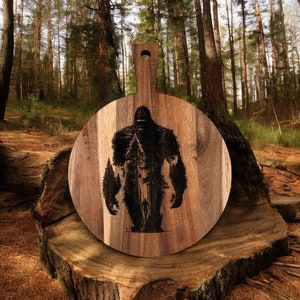 May include: A round wooden cutting board with a handle. The board has a laser-etched design of Bigfoot standing in a forest.