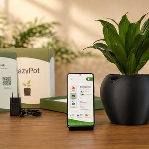 Op de afbeelding: Een zwarte, ronde slimme plantenpot met een groene plant, naast een smartphone met een app-interface, een LazyPot-doos, een gedrukte handleiding en een zwarte stroomadapter met snoer op een houten ondergrond.