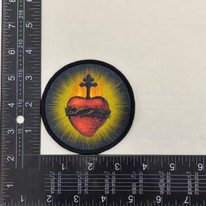May include: Round patch featuring a red heart with a crown of thorns, a cross, and yellow rays on a black background. The heart is centered, with a black border. Religious themed.