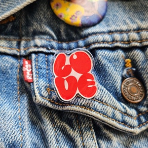 Peut inclure: Une épingle en plastique rouge avec le mot "LOVE" en lettres bulles. L'épingle est fixée à une veste en jean.