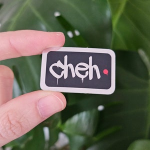 Puede incluir: Una pegatina rectangular con esquinas redondeadas. La pegatina tiene un fondo negro con la palabra "cheh" en letras blancas de estilo grafiti. Un pequeño punto rojo está en el lado derecho. La pegatina está en la mano de una persona.