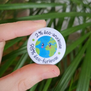 Könnte beinhalten: Ein runder, weißer Button mit dem Text "2% eco-anxieuse, 98% eco-furieuse", der eine Cartoon-Erde mit traurigem Gesicht umgibt. Die Erde ist blau und grün, mit grünen Punkten am Rand.