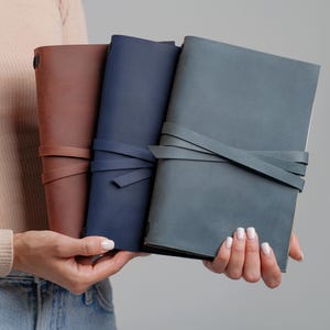 May include: Three leather journals in shades of brown, navy blue, and gray are held in front of a neutral background. Each journal has a wraparound strap closure. The journals are a classic design, suitable for writing or sketching.