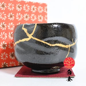 Könnte beinhalten: Eine schwarz glasierte Keramikschale mit goldfarbenen Kintsugi-Reparaturlinien auf der Oberfläche. Die Schale steht auf einem kleinen quadratischen weinroten Untersetzer neben einer roten Schachtel mit weißem Spiralmuster.