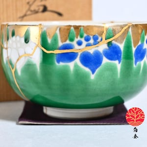 May include: A ceramic bowl with a green and white floral design, accented with blue heart-shaped flowers. The bowl features gold-coloured kintsugi repair lines. The bowl is on a purple mat, with a wooden box in the background.
