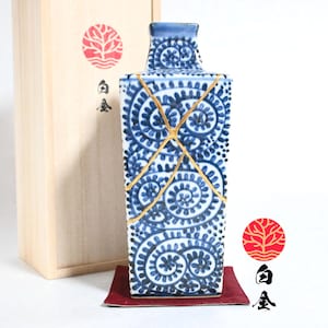 May include: A square ceramic bottle featuring a blue and white spiral pattern with gold-colored kintsugi repair lines. The bottle is accompanied by a light-colored wooden storage box marked with a red circular tree logo and black Japanese text.