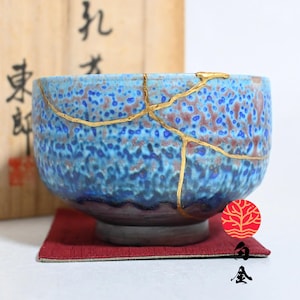 Può includere: Una ciotola da tè in ceramica caratterizzata da uno smalto blu e viola maculato con riparazioni in lacca dorata sulla superficie. La ciotola poggia su un piccolo quadrato di tessuto rosso, posizionato davanti a una scatola di legno chiaro.