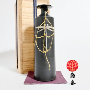 Può includere: Una bottiglia in ceramica nera, alta e cilindrica, caratterizzata da un motivo di riparazione kintsugi dorato e da un disegno di libellula bianca. La bottiglia poggia su un piccolo tappetino quadrato viola accanto a una scatola in legno chiaro.