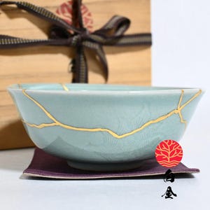 May include: A light blue ceramic bowl featuring gold-coloured kintsugi repair lines across its surface. The bowl rests on a dark purple fabric square, positioned in front of a light brown wooden box tied with a dark brown ribbon.