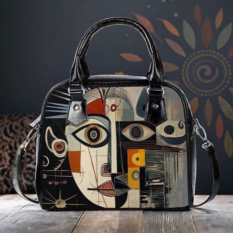 May include: Black handbag with a unique abstract face design. The bag features a black handle and a detachable shoulder strap. The artwork incorporates geometric shapes and a palette of black, white, orange, and yellow.