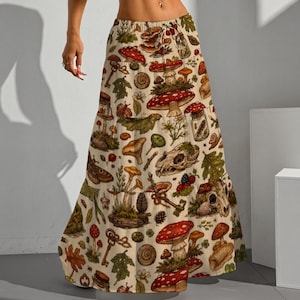 Puede incluir: Falda larga color crema con un estampado caprichoso de setas y temática forestal. El diseño incluye setas de sombrero rojo, llaves, hojas y calaveras de animales. La falda tiene una silueta fluida y escalonada.