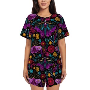 May include: A two-piece pajama set featuring a vibrant, all-over print of purple moths, flowers, and botanical elements on a black background. The short-sleeved top has a button-up neckline, and the shorts complete the set. The design is colorful and detailed.