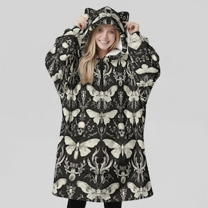 May include: Black hooded blanket with a white butterfly and skull pattern. The blanket has a zipper closure and cat ear details on the hood. The interior lining is white.