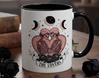 May include: White ceramic mug with a black interior and handle. The mug features a design of two skeletons embracing within a heart, surrounded by crescent moons and stars. The words "THE LOVERS" are printed on a banner below the heart.