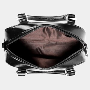 May include: Open view of a black, glossy handbag with a zipper closure. The bag has two handles and silver-colored hardware. The interior is lined with a brown fabric.