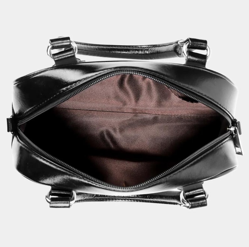 May include: Open view of a black, glossy handbag with a zipper closure. The bag has two handles and silver-colored hardware. The interior is lined with a brown fabric.