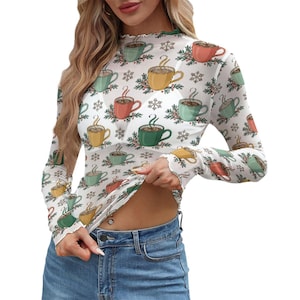Pode incluir: Uma blusa branca de manga comprida com gola alta e um padrão repetido de xícaras de café coloridas, flocos de neve e galhos. A blusa tem uma borda com babados no pescoço e punhos. A modelo está usando jeans azuis.
