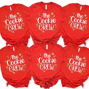 May include: Six red short-sleeved t-shirts arranged in two rows. Each shirt features the white text The Cookie Crew in a script font, surrounded by small colorful illustrations of gingerbread people, cookies, candy canes, and star shapes.