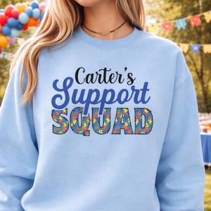 May include: A light blue sweatshirt with the text "Carter's Support Squad" in a mix of fonts and colors. The word "Squad" is filled with a colorful pattern. The sweatshirt is a crew neck style.