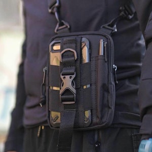 Puede incluir: Una funda de teléfono táctica negra con una correa y un porta bolígrafos. La funda tiene una cremallera y una hebilla.