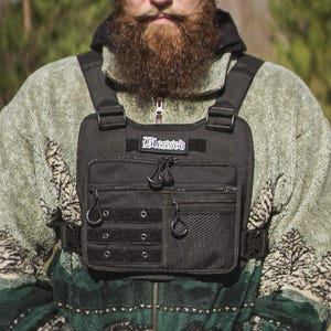 Peut inclure: Un harnais de poitrine noir comprenant une pochette rectangulaire avec une poche zippée frontale, un compartiment en filet et un écusson portant le mot Blessed. Le harnais possède des bretelles réglables avec boucles sur une veste polaire verte.