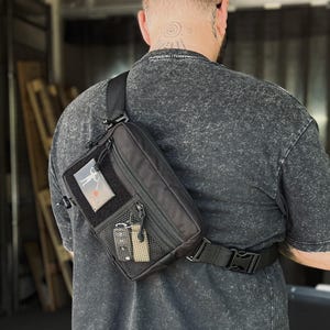 May include: A black crossbody bag with a zippered main compartment, a front mesh pocket, and a rectangular patch area. The bag features a black adjustable strap with a side-release buckle and is worn across the back over a dark gray t-shirt.