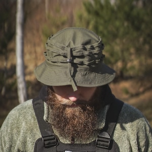 May include: Olive green bucket hat with a structured crown and a chin strap. The hat is paired with a black tactical vest featuring the word "Blessed" on a patch. The image has a natural, outdoor setting.