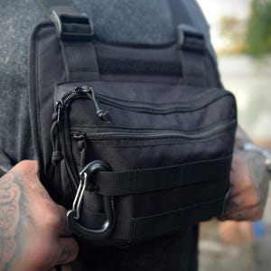 May include: Black chest rig with multiple compartments and adjustable straps. Features a zippered main pocket, a carabiner clip, and MOLLE webbing for attaching additional gear. The rig is designed to be worn over the chest.