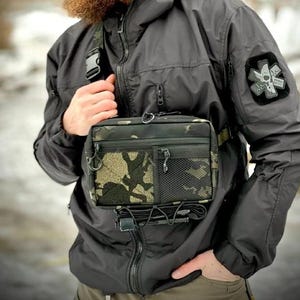 Puede incluir: Una bolsa de pecho negra con un patrón de camuflaje en la parte delantera. La bolsa tiene múltiples compartimentos, incluyendo un bolsillo de malla y un bolsillo con cremallera. La bolsa se lleva cruzada sobre el pecho con una correa ajustable. También se ve una chaqueta negra con un parche.