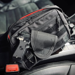 May include: Black tactical fanny pack with a red zipper lining and a handgun. The pack has multiple compartments, Velcro straps, and a holster. The handgun is black and silver.