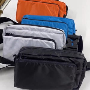 May include: A selection of rectangular fanny packs in orange, blue, white, and black. Each pack features a front pocket with a zip and a black strap with a buckle. The packs are made of a smooth, possibly nylon, material.